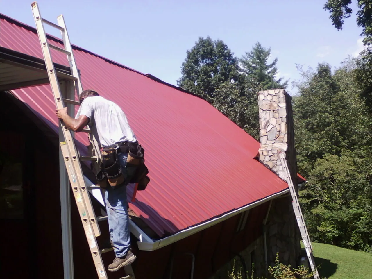 Roofer on a ladder beside a red metal roof and stone chimney — Soffit & Fascia Repair by Roofing Brothers in Millerstown